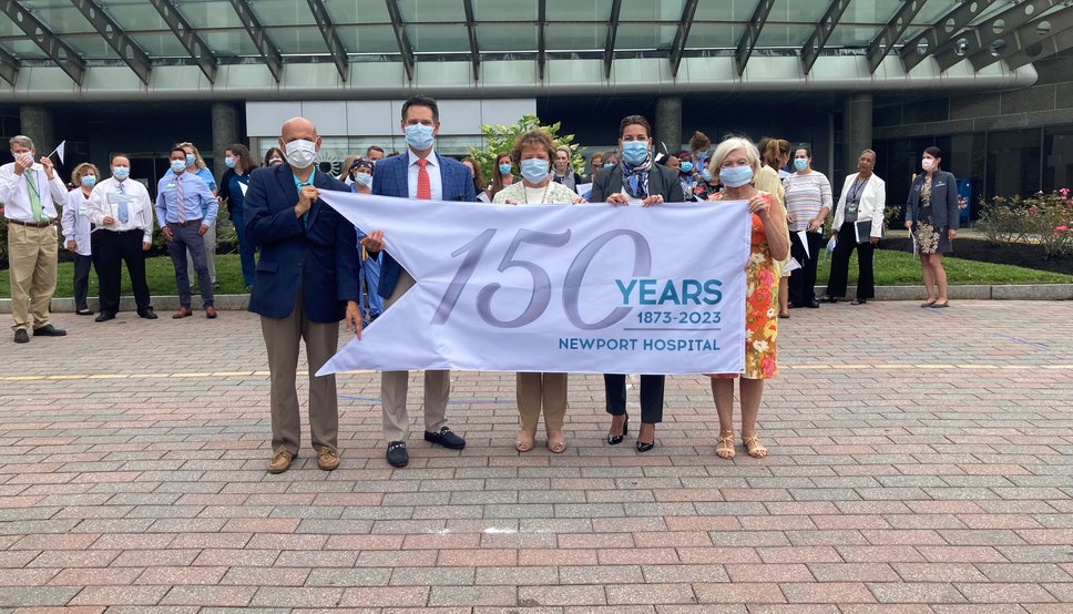 photo of hospital leaders displaying a flag that says Newport Hospital 150 years old