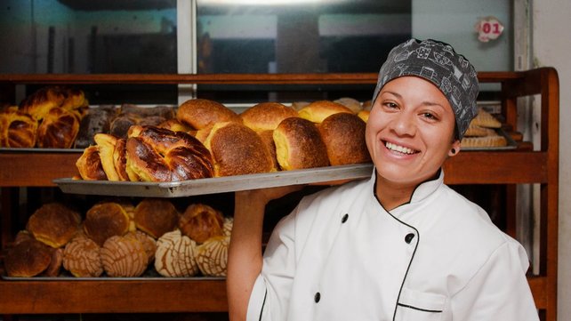 Woman in chef coat balancing tray of pastries on shoulder and smiling