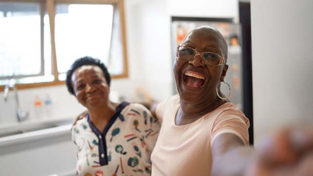 Two women in kitchen laughing and gesturing to camera 