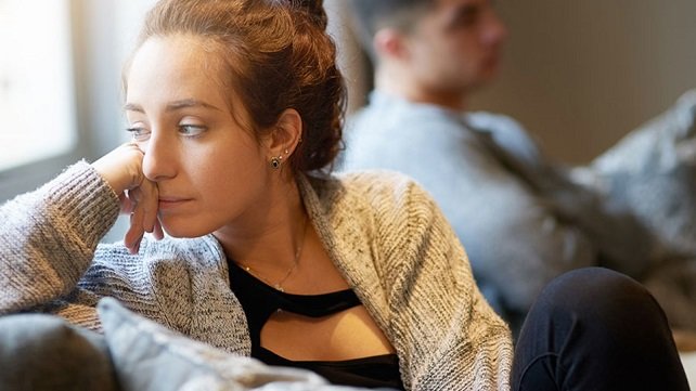 Young woman with auburn hair and wearing a sweater sitting on a couch and resting her chin in her hand while looking to the side