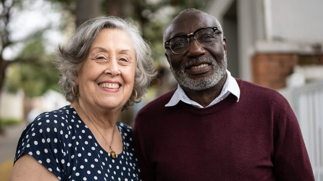Close up of an older woman and man in front of their house, smiling.
