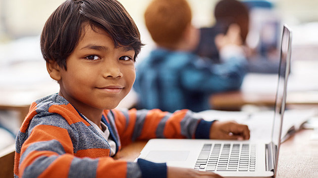 Smiling young boy, wearing a striped shirt, sitting at a desk with a laptop