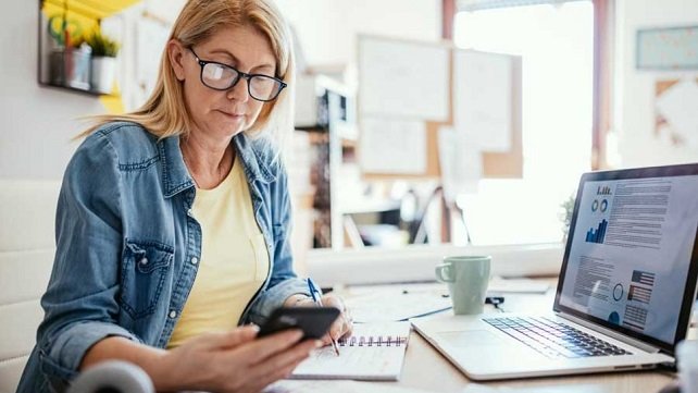 Middle-aged woman with blonde hair and glasses in her kitchen looking at her cell phone and working on her laptop