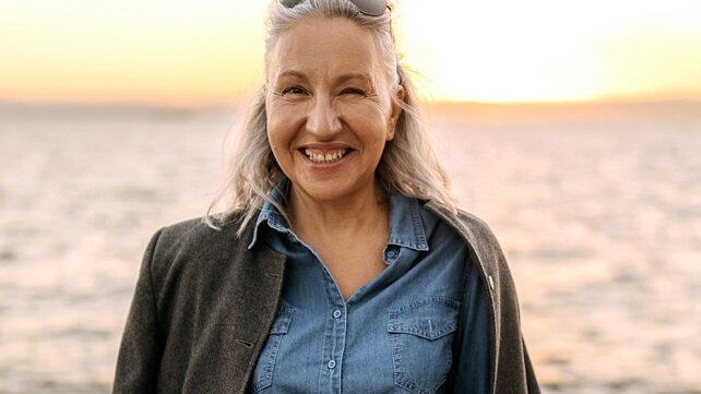 Smiling older woman standing on a beach with the water behind her