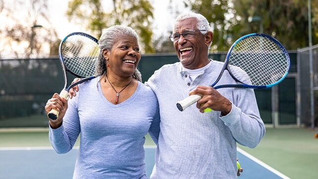 An older couple smiles and laughs while holding tennis rackets on a tennis court.