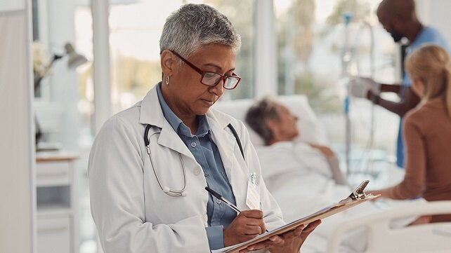 A doctor taking notes while a family member visits a patient in the background.
