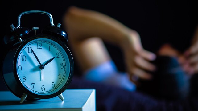 Photo of an alarm clock (in the foreground) with current time of 1:55 a.m. on a bedside table next to a bed with a restless person in it.
