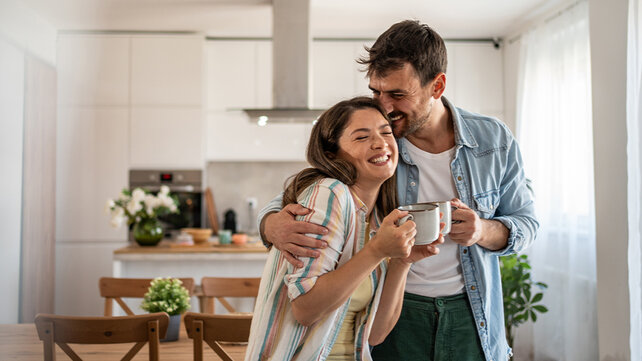 A man hugs a woman. Each of them is holding a mug of coffee.