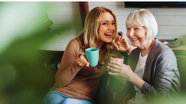 An adult woman and her mother, drinking tea, on a porch swing on a summer day.