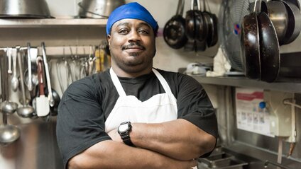 Man in restaurant kitchen wearing apron and smiling with arms crossed
